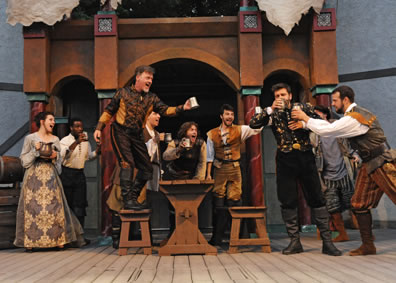 Iago stands on a bench next to a table with tankard held out, standing next to the bench on the opposite side is Cassio in black drinking, as Montano holds a tankard up to him as well. Ranged across the background are men with tankards and a woman with a pitcher (to the left) all cheering, and in the background a three-arch wood doorway with red marble-looking columns and ragged sheets hanging down at the top.